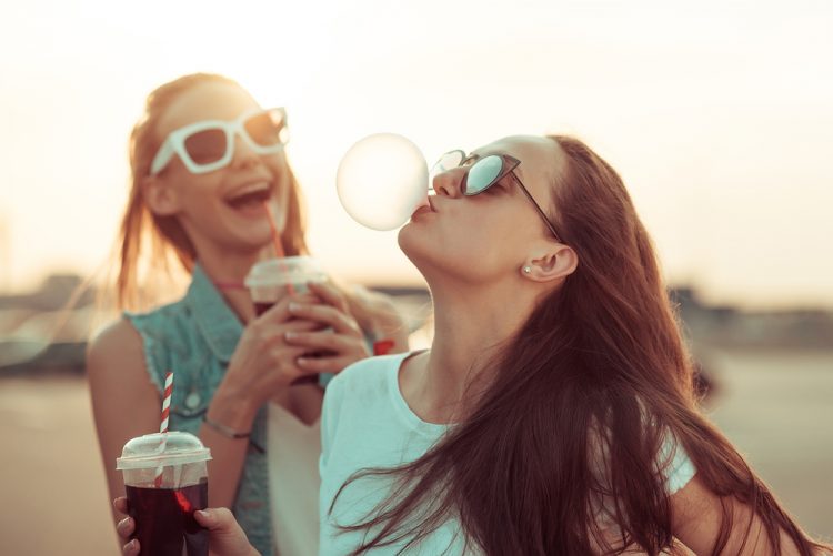 Two teen girls at dusk, laughing, sipping drinks, and blowing a bubblegum bubble